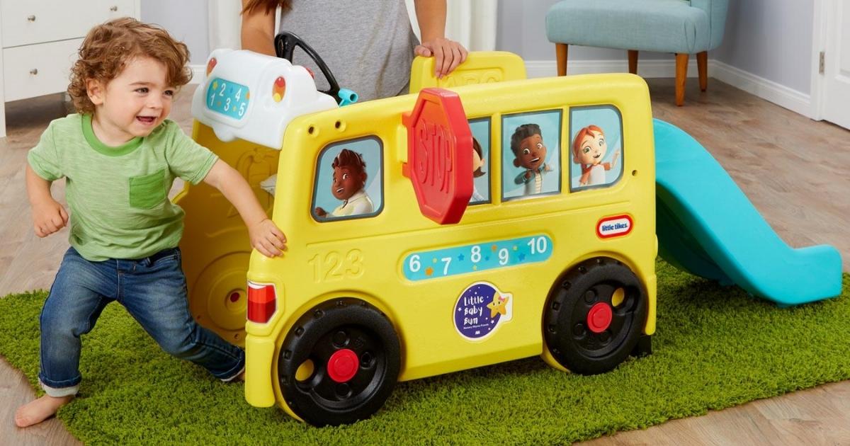 laughing toddler playing next to the Little Tikes Little Baby Bum Bus playset while on green carpeting