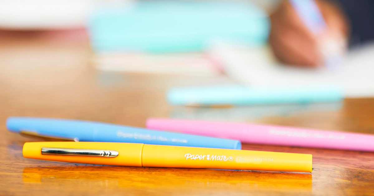 a few colorful pens in focus on a desk with a blurred background showing someone writing