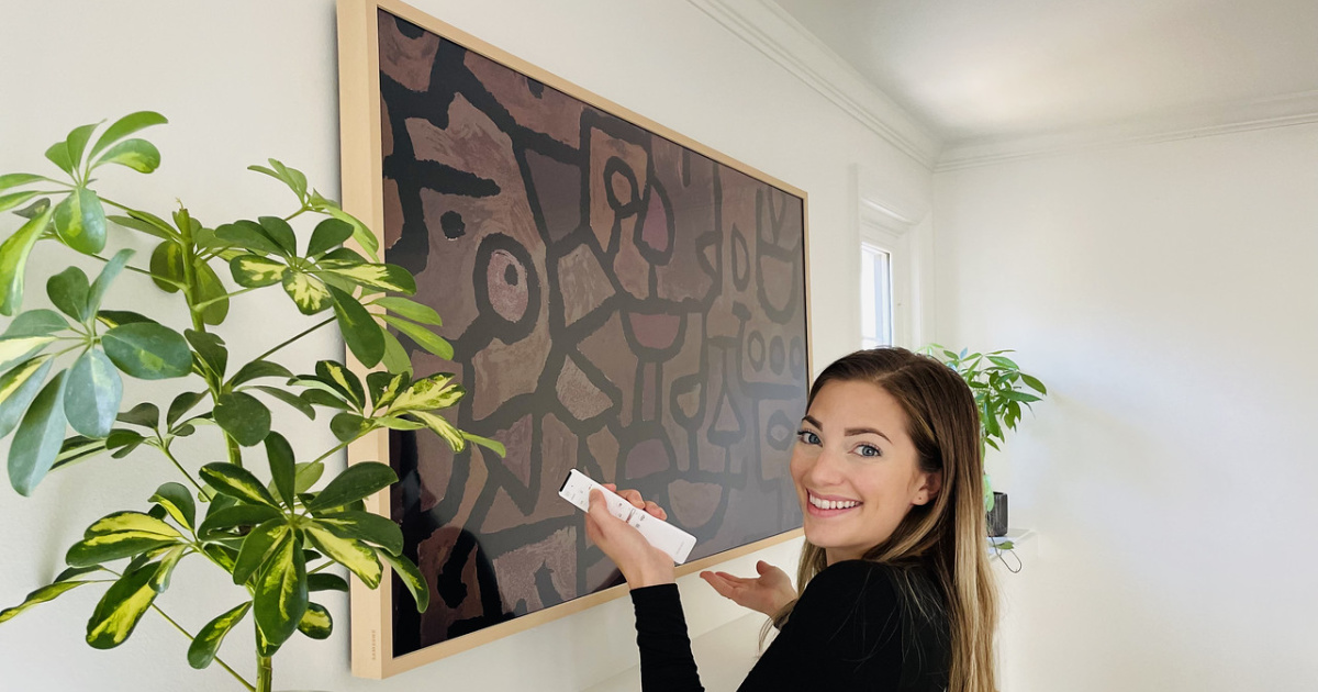 woman standing in front of a framed piece of art and plant, holding a remote