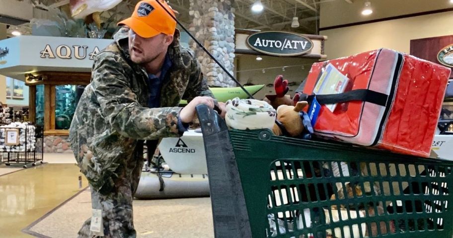 man wearing camo hunting clothing and an orange hat pushing a cart full of items in Bass Pro shop