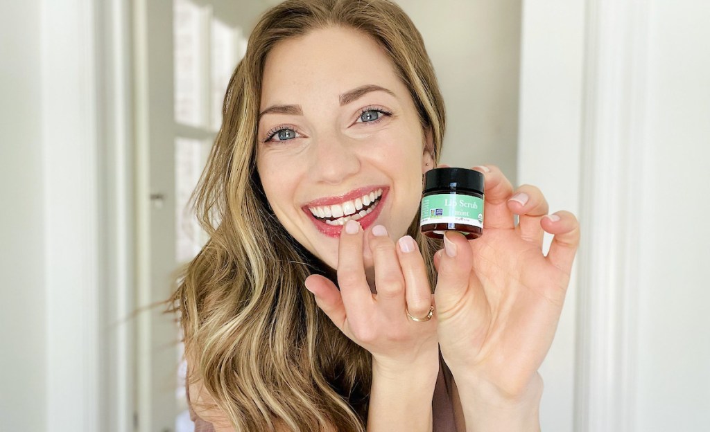 woman holding lip scrub while applying it on lips smiling