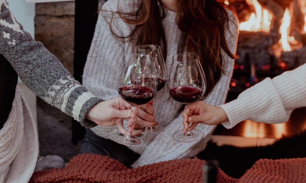 three people holding red wine glasses together in front of fireplace