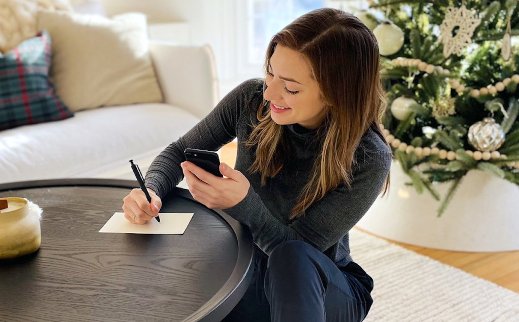 woman writing on paper while holding phone with hallmark send a card program