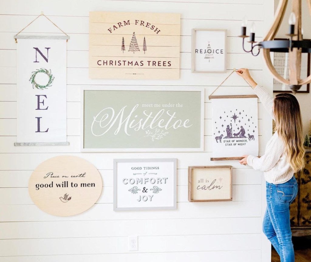 woman hanging christmas sign on white wall with various holiday wood signs