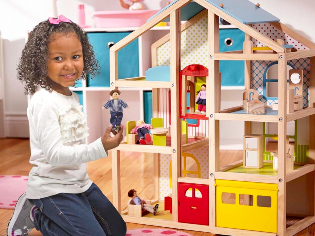 girl kneeling next to a wooden dollhouse holding a doll in her hands