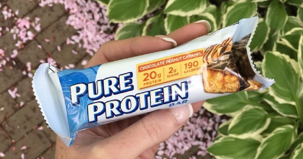 Pure Protein Bars 12-Count Chocolate Peanut Caramel in womans hands in front of greenery