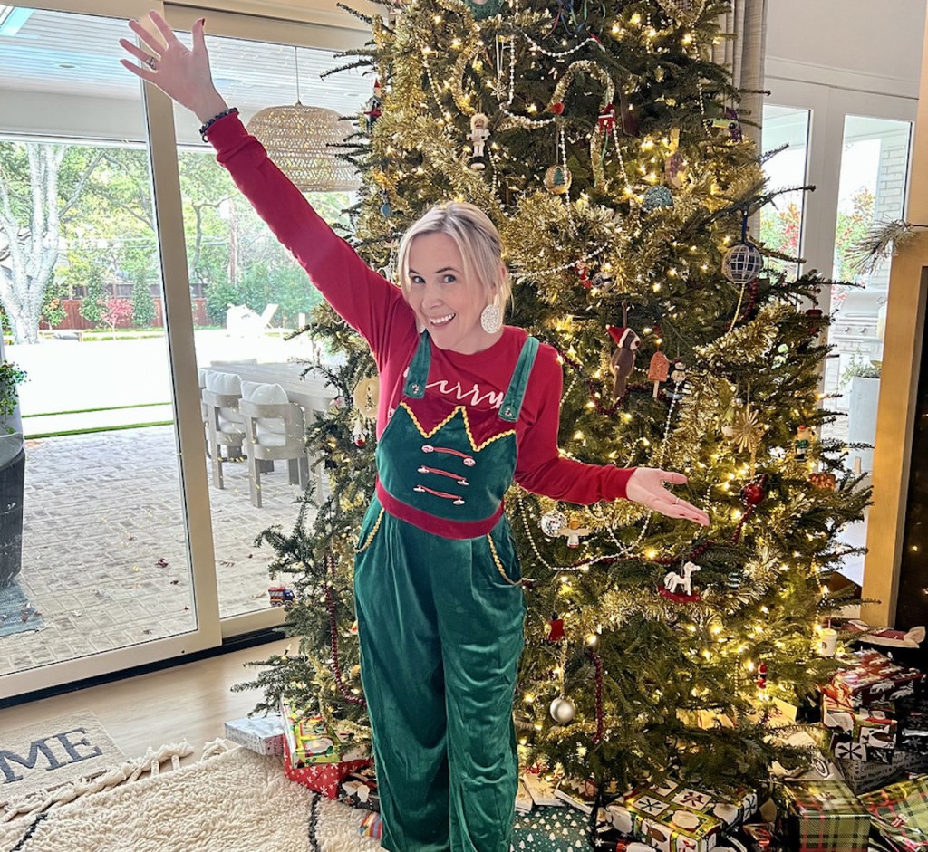 woman wearing christmas overall in front of lit christmas tree