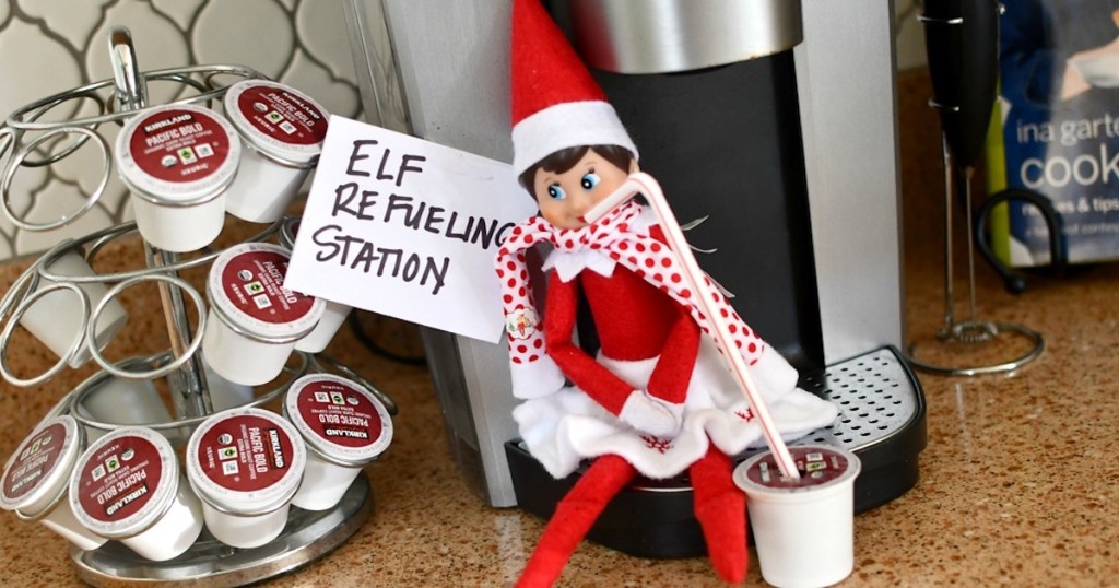 elf on the shelf sipping straw from k cup coffee on kitchen counter