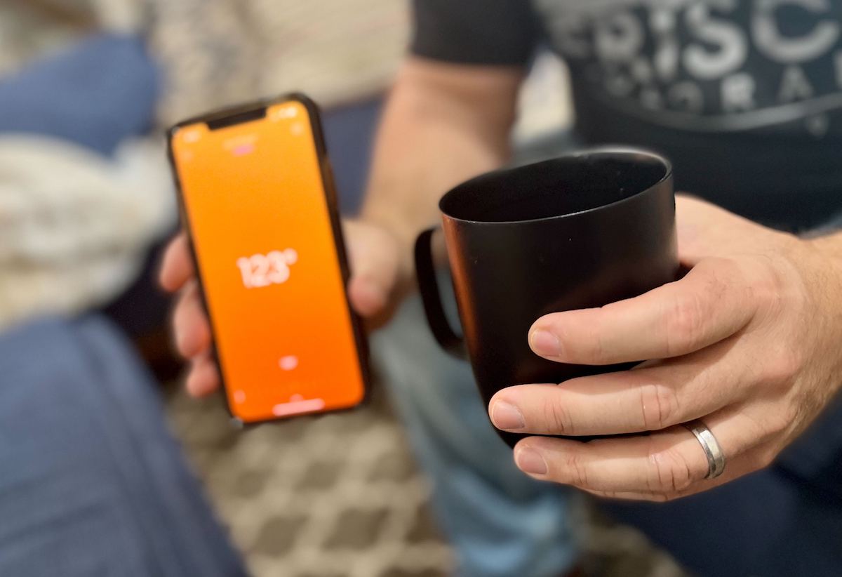 man holding black ember mug with phone showing orange screen with temperature reading
