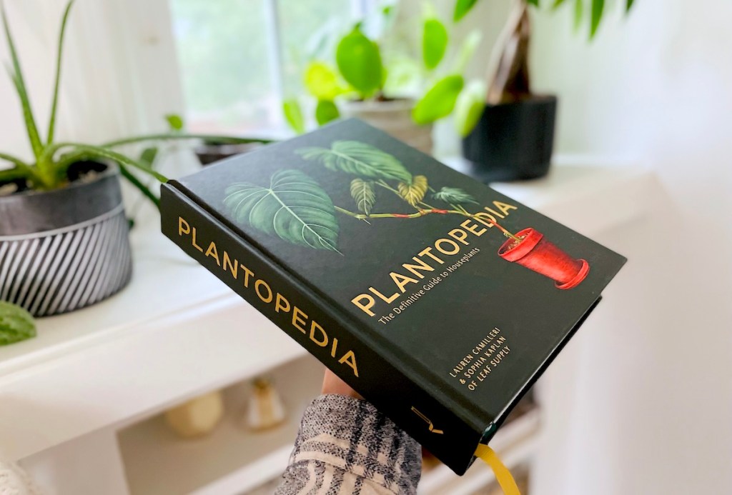 arm holding out black plantopedia book in front of various green plants on bookshelf