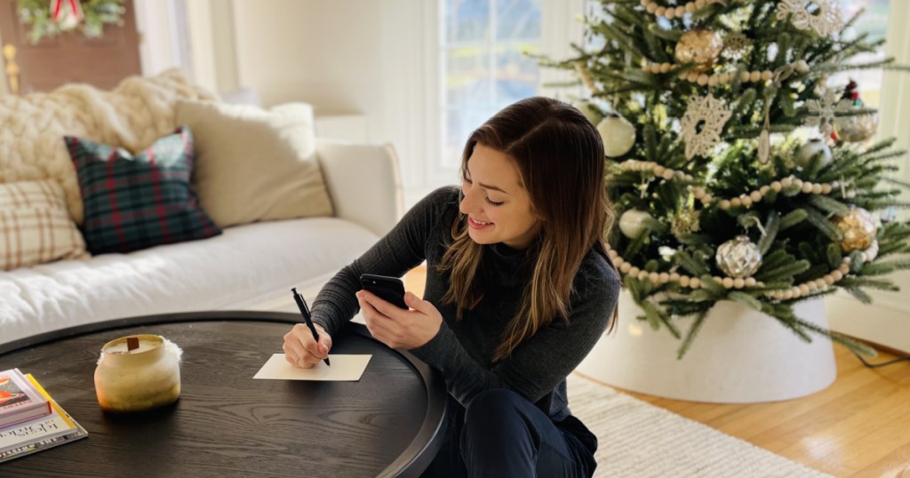 woman sitting on the floor at a coffee table writing a card while holding her phone with a christmas tree in the background