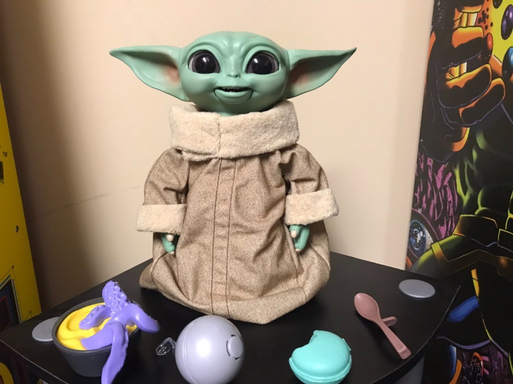 Baby Yoda Doll in home sitting next to Yoda's toys