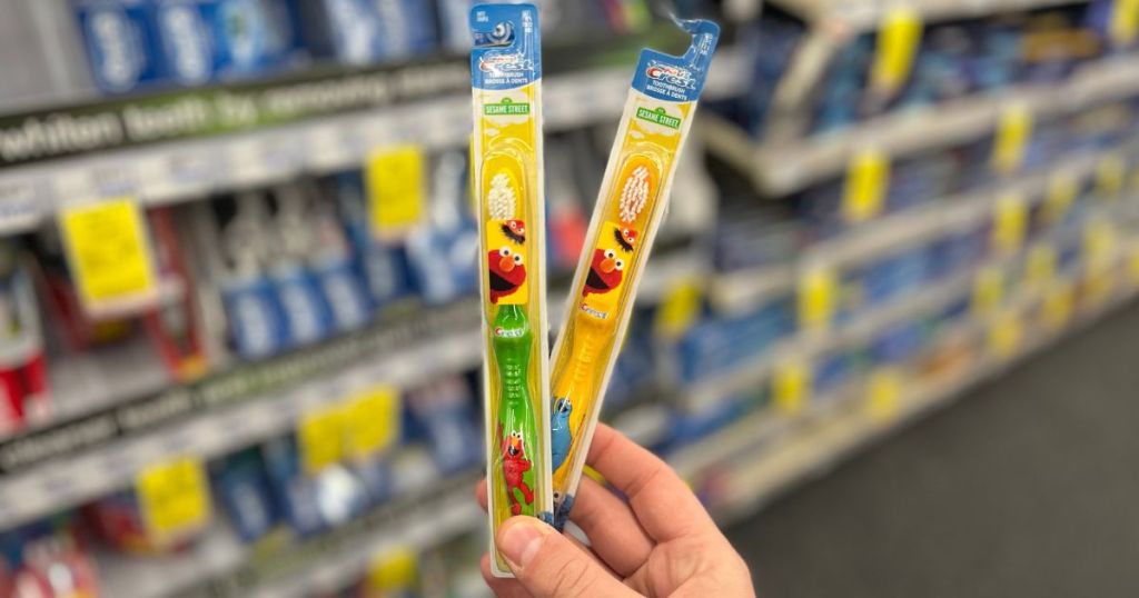 hand holding 2 Crest Kid's Sesame Street Soft Bristles Toothbrushes