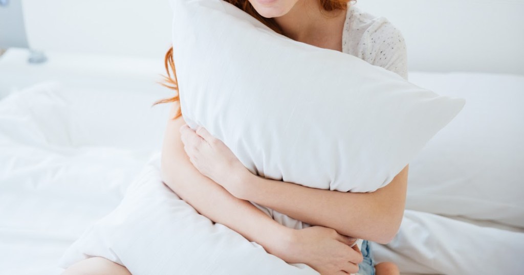 woman sitting on bed hugging large white bed pillow