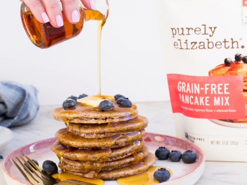 woman pouring syrup on stack of pancakes and bag of grain-free pancake mix