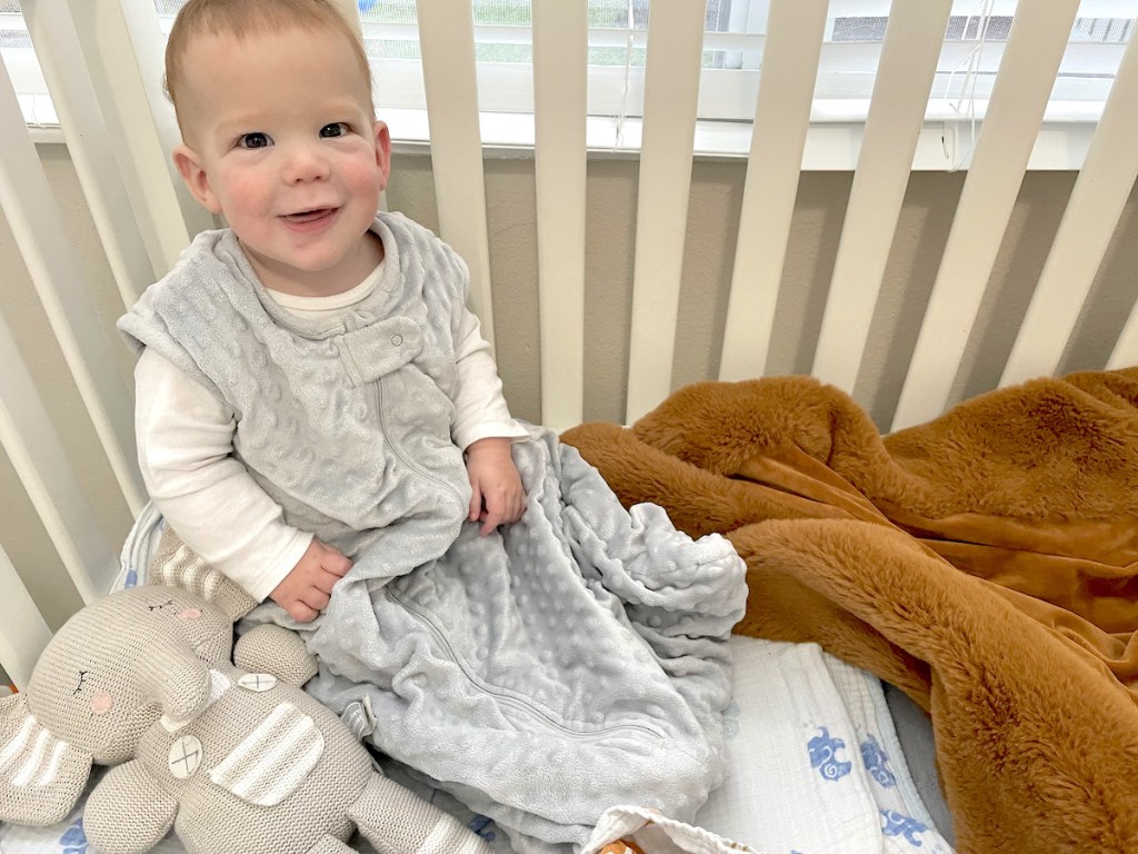 baby boy with gray mink sleepsack sitting in crib smiling