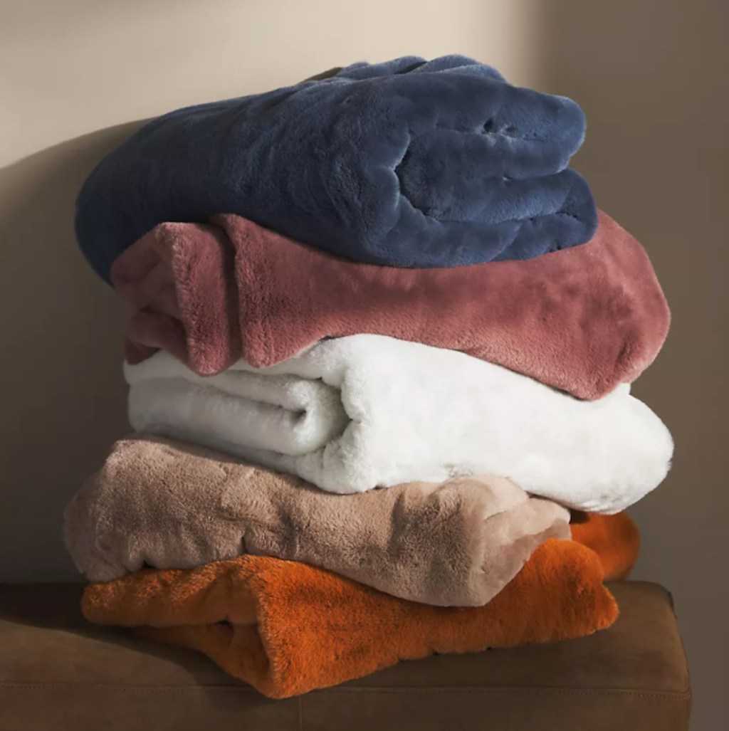 stack of solid colored fur throw blankets on brown ottoman in front of brown wall