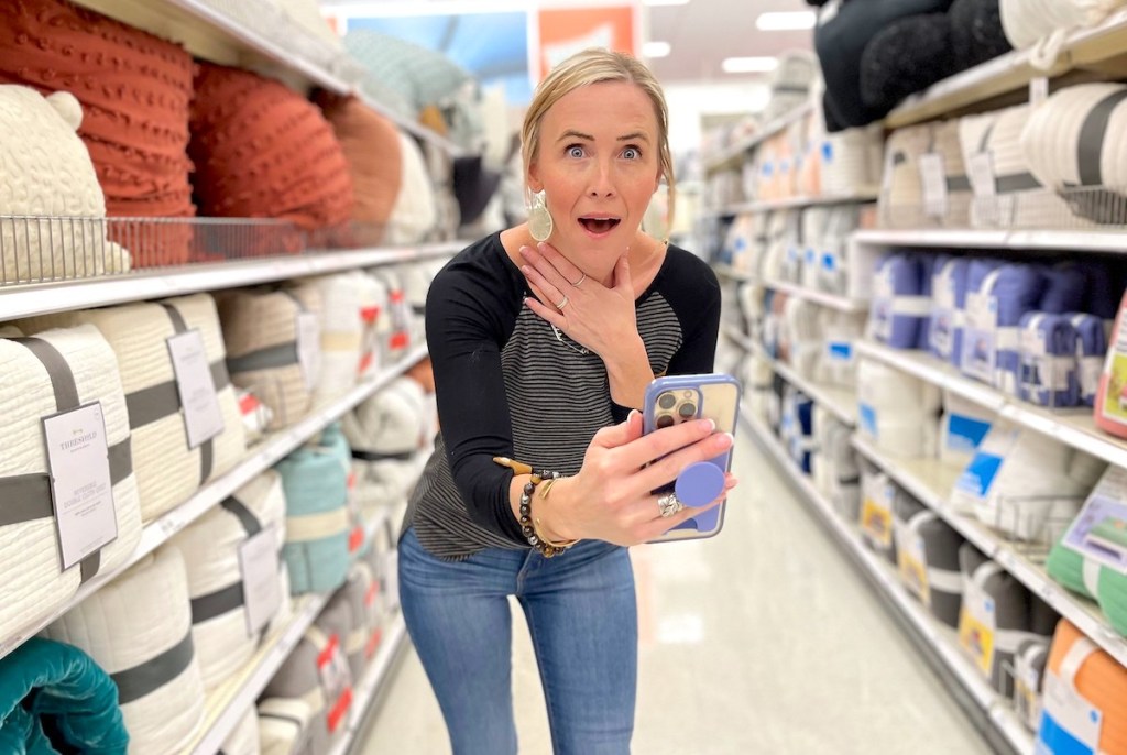 woman with hand on chest holding out phone in store bedding aisle