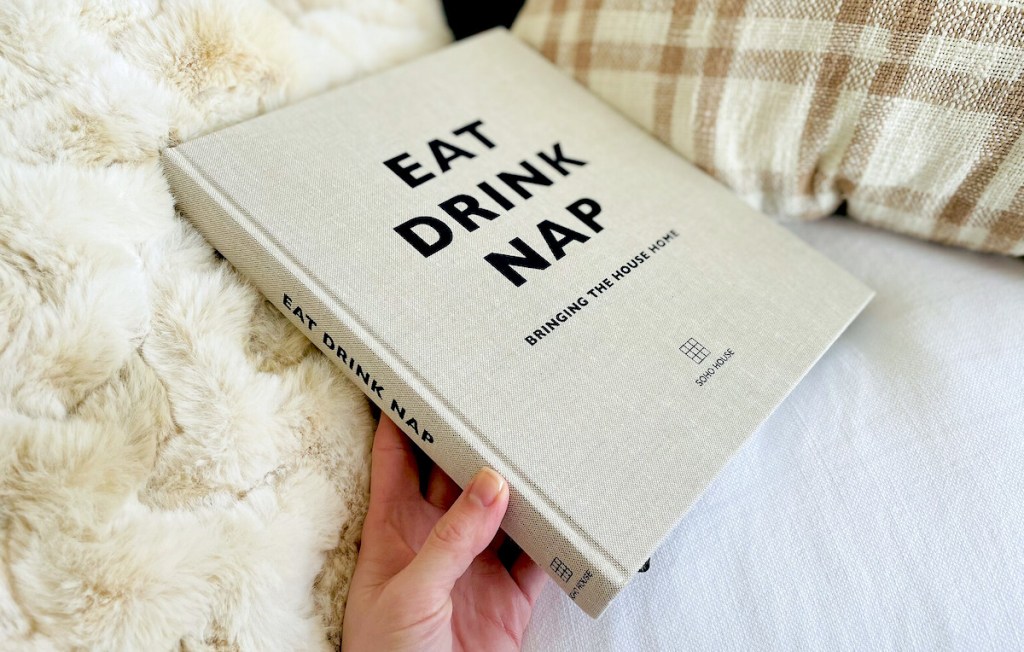 hand holding eat drink nap linen hardcover coffee table book on white couch with blanket and plaid pillow