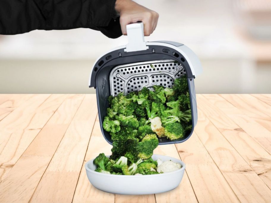 hand pouring broccoli florets from a white air fryer basket onto a plate
