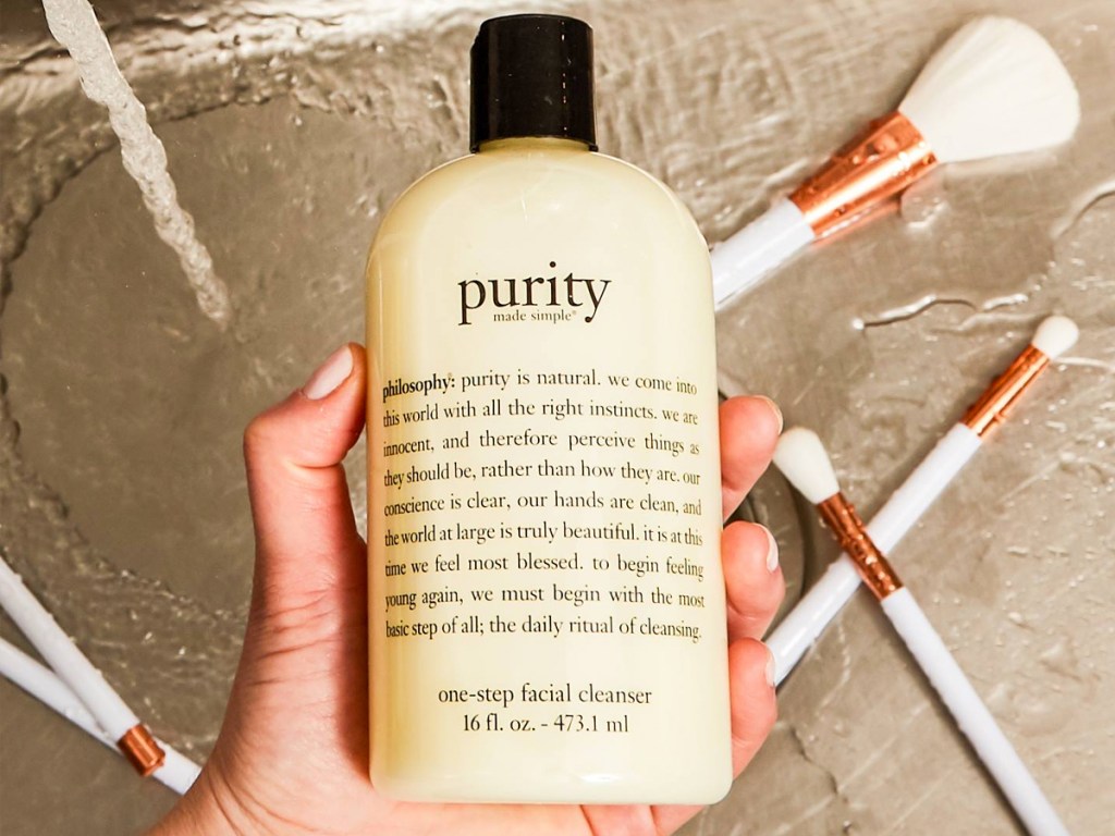 hand holding up a Philosophy Purity Made Simple One-Step Facial Cleanser 16oz Bottle in sink with makeup brushes