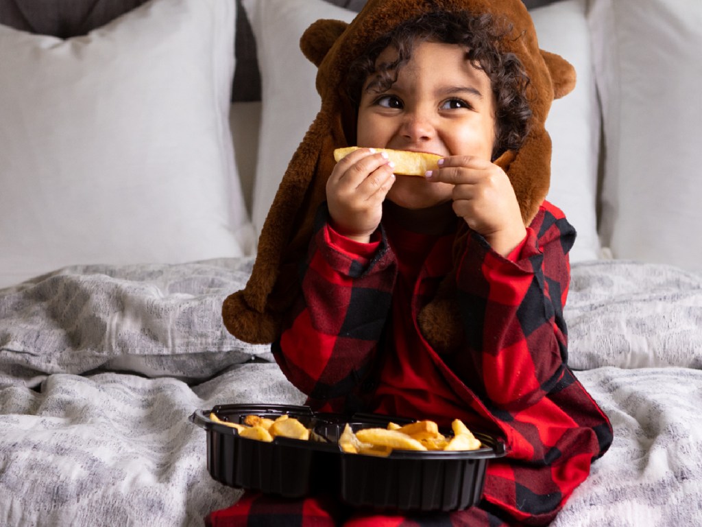 boy sitting on bed in pajamas eating fries from takeout container