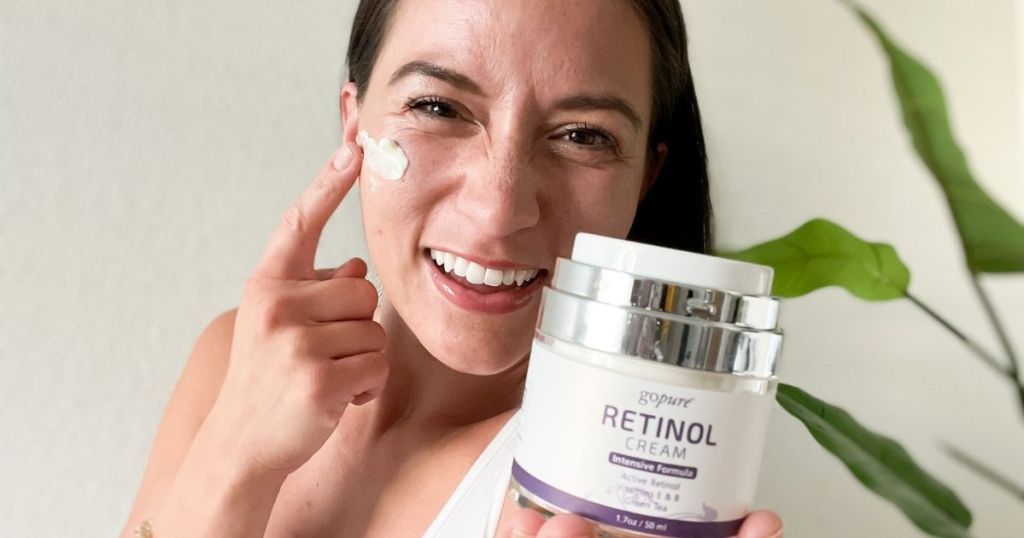 woman holding up Go Pure Retinol Cream with one hand and rubbing cream on face with other hand