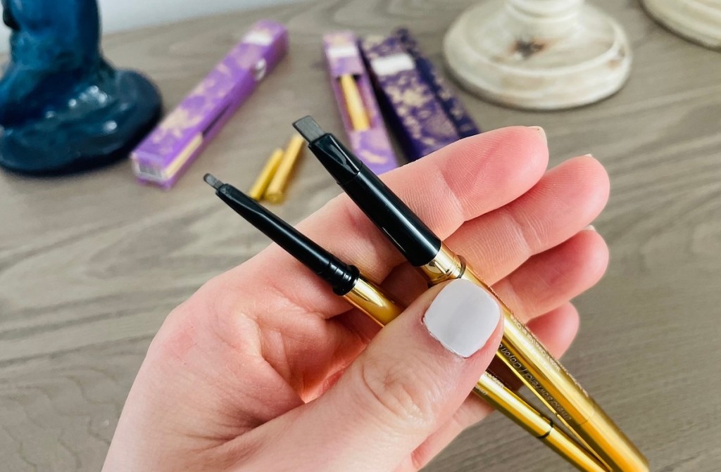 hand holding two gold and brown eyebrow pencils