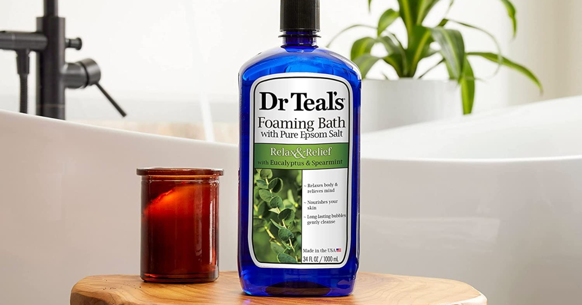 Blue bottle of Dr. Teals foaming bath on a wooden stool with a candle, in a bathroom. A bathtub is in the background.