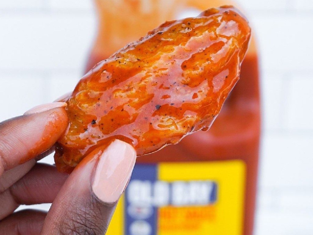 hand holding chicken wing in front of large bottle of hot sauce