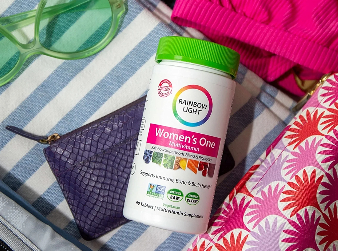 Rainbow Light Women's Vitamin jar laying by towel, green sunglasses and purple cosmetic bag
