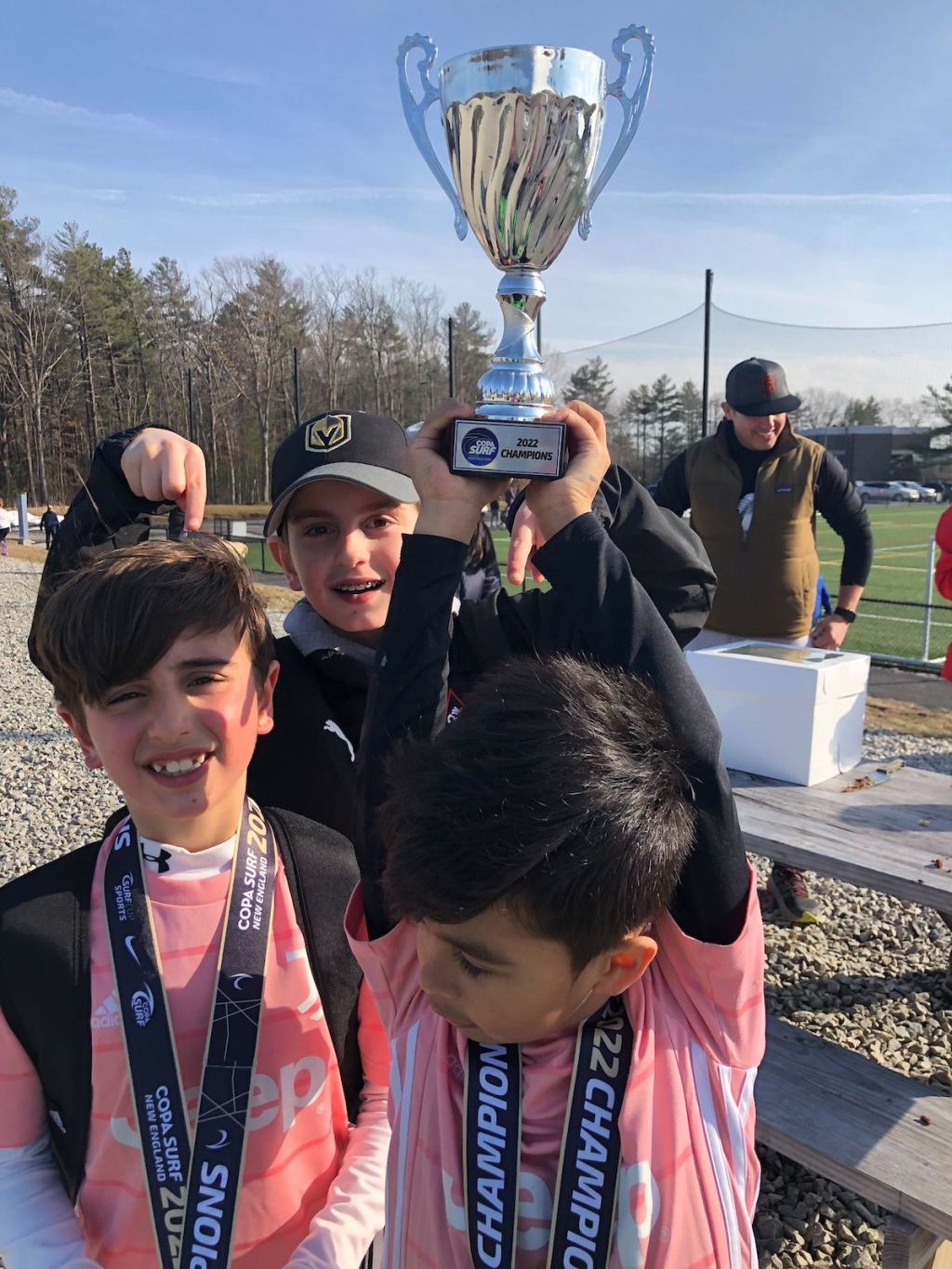three boys holding medals and soccer trophy smiling outside