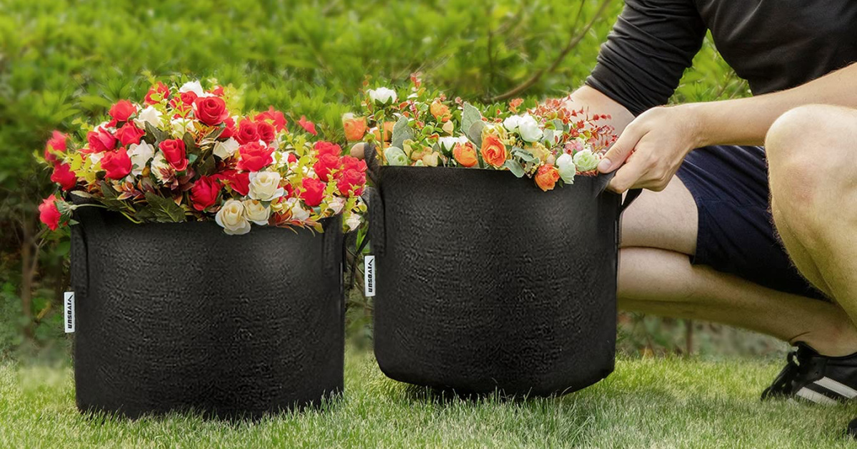 two black grow bags filled with various flowers being tended by a man in shorts and sneakers