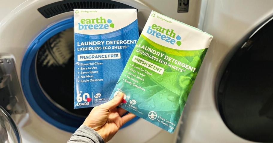 Hand holding two packs of Earth Breeze laundry sheets in front of a washing machine