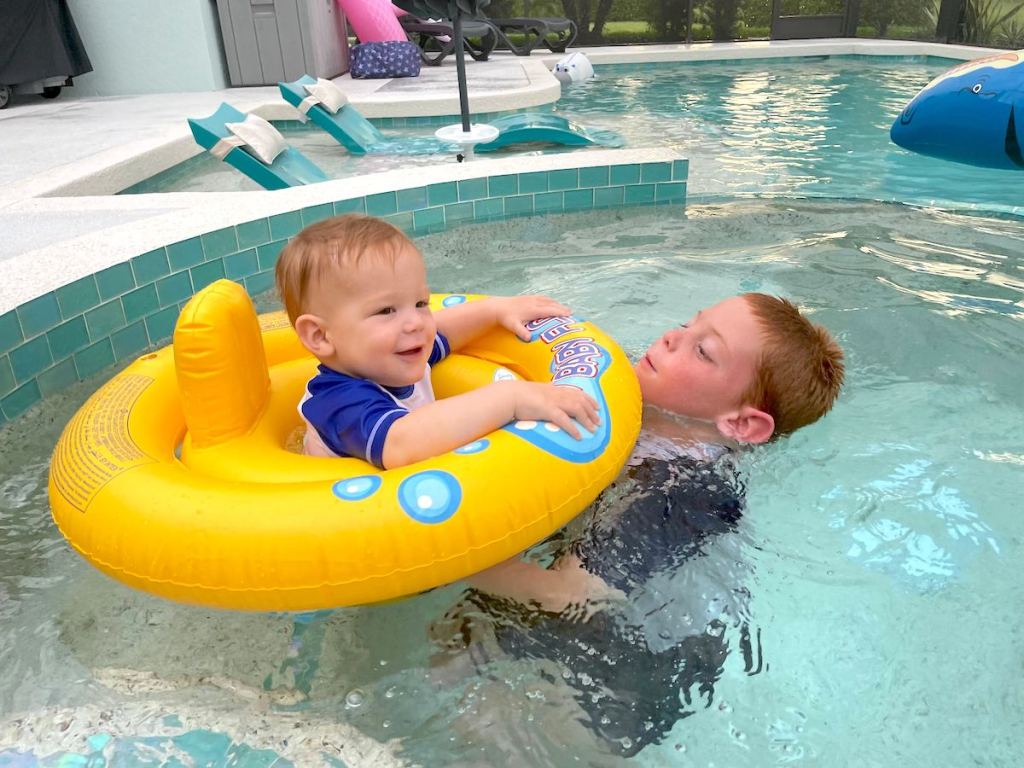baby in yellow pool float with big kid swimming in in ground pool