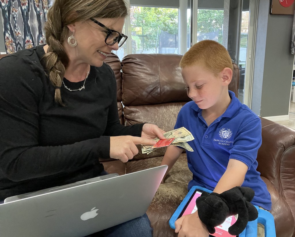 woman holding money and debit card on couch with laptop smiling at son