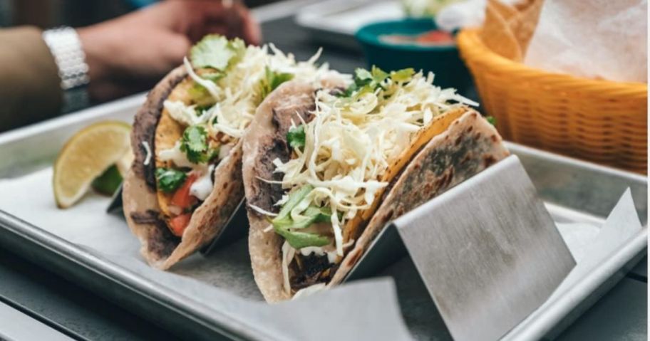 tacos sitting in a stainless steel taco holder stand on a platter on a table with other food