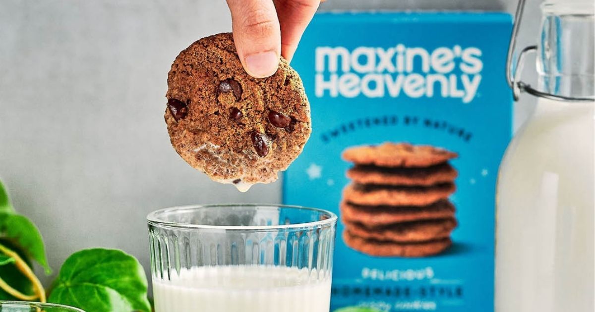 hand dipping cookie into glass of milk with Maxine’s Heavenly Cookies package in background