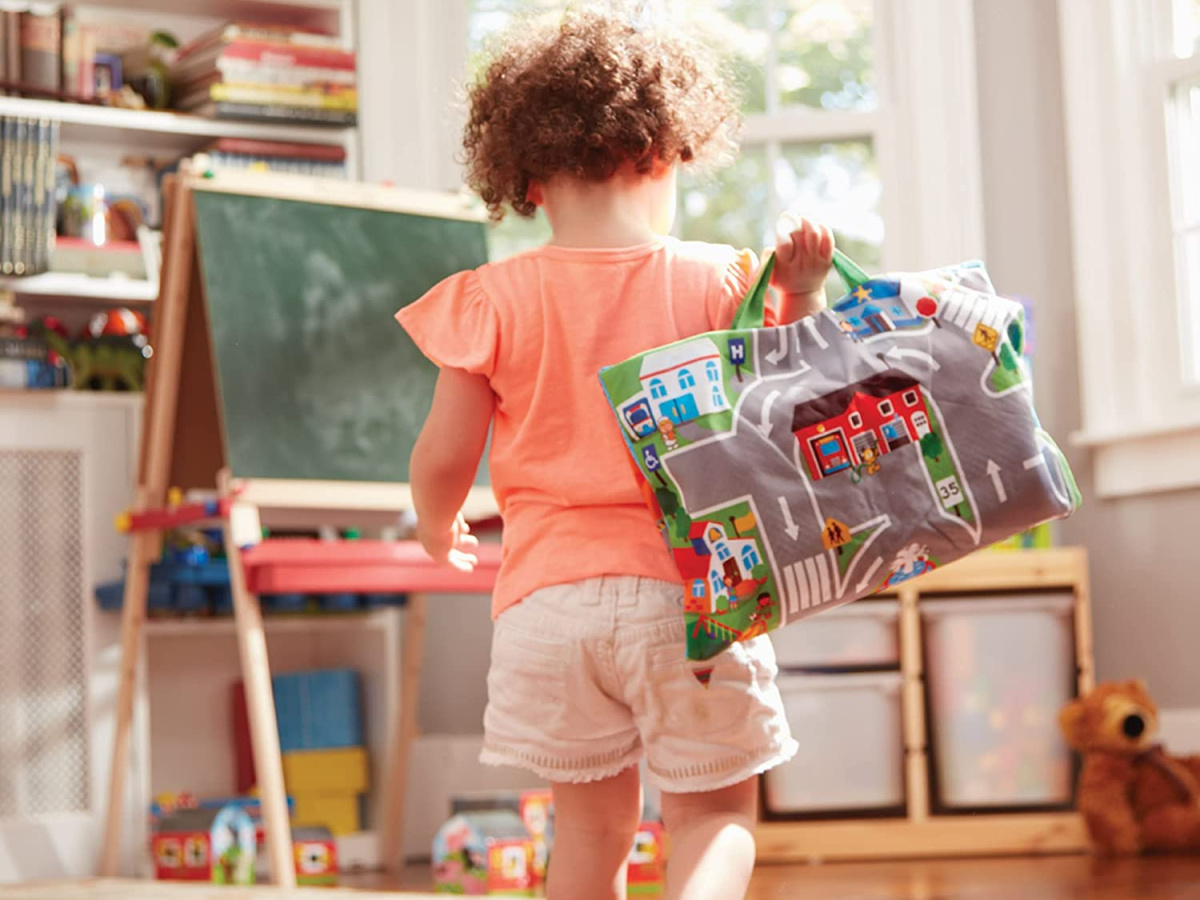 young girl facing away from the camera carrying the melissa and doug take along town mat tote in a playroom