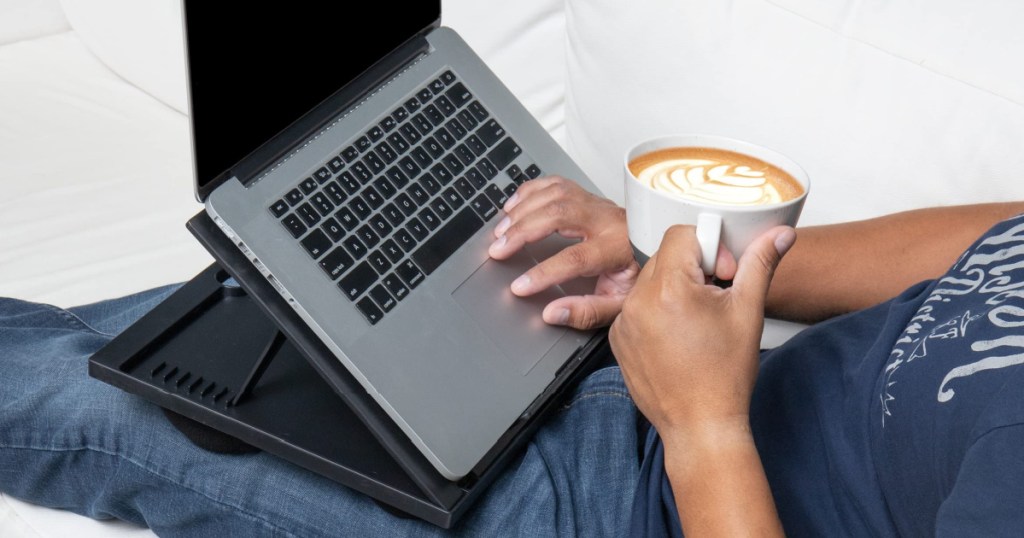 man holding latte and using laptop sitting on lap desk