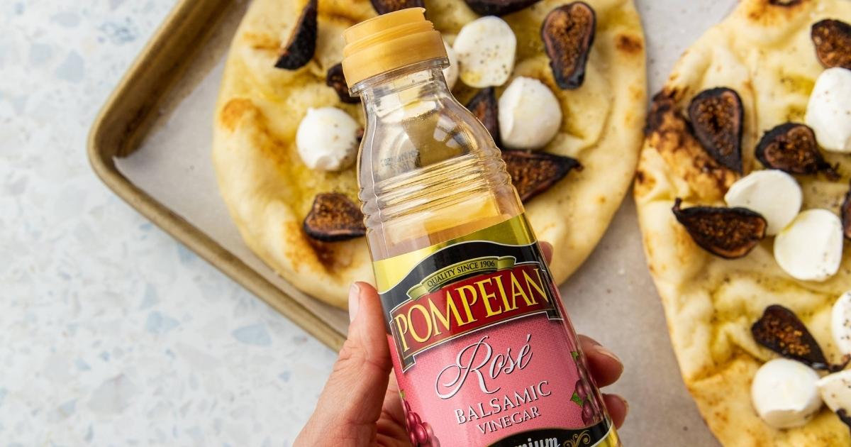 hand holding a bottle of Pompeian Vinegar above two mozzarella and fig pizzas on a sheet pan