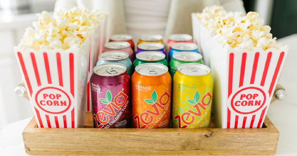 woman holding a tray with cans of Zevia Soda between cartons of popcorn