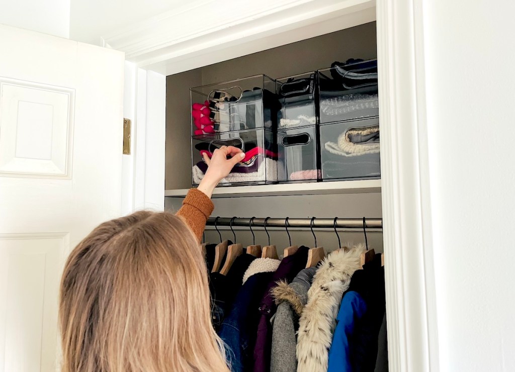 woman reaching into coat closet to grab clear bins with winter gear inside