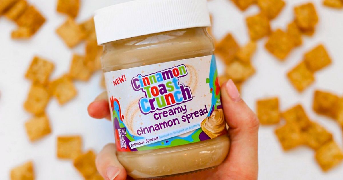 a hand holding a jar of Cinnamon toast crunch creamy spread, with a background of scattered cinnamon toast crunch cereal
