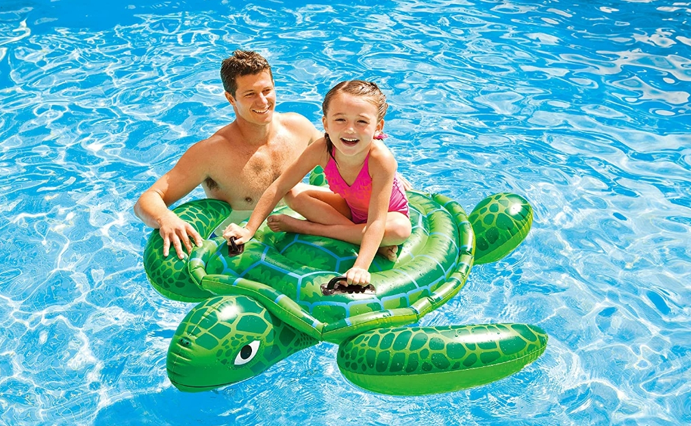 Girl riding on a turtle inflatable pool float in a pool with a man standing next to her