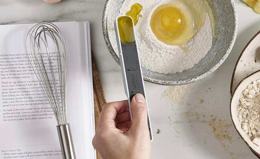Hand holding measuring spoon with olive oil over baking bowl on the counter top with recipe book