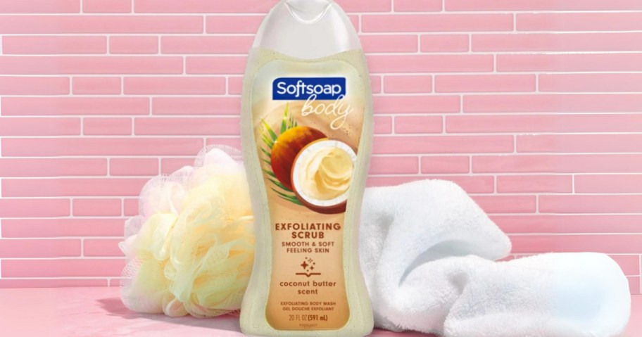 a bottle of Softsoap Exfoliating Body Wash in Coconut Butter Scrub next to a mesh sponge and towel, pink subway tiles behind them