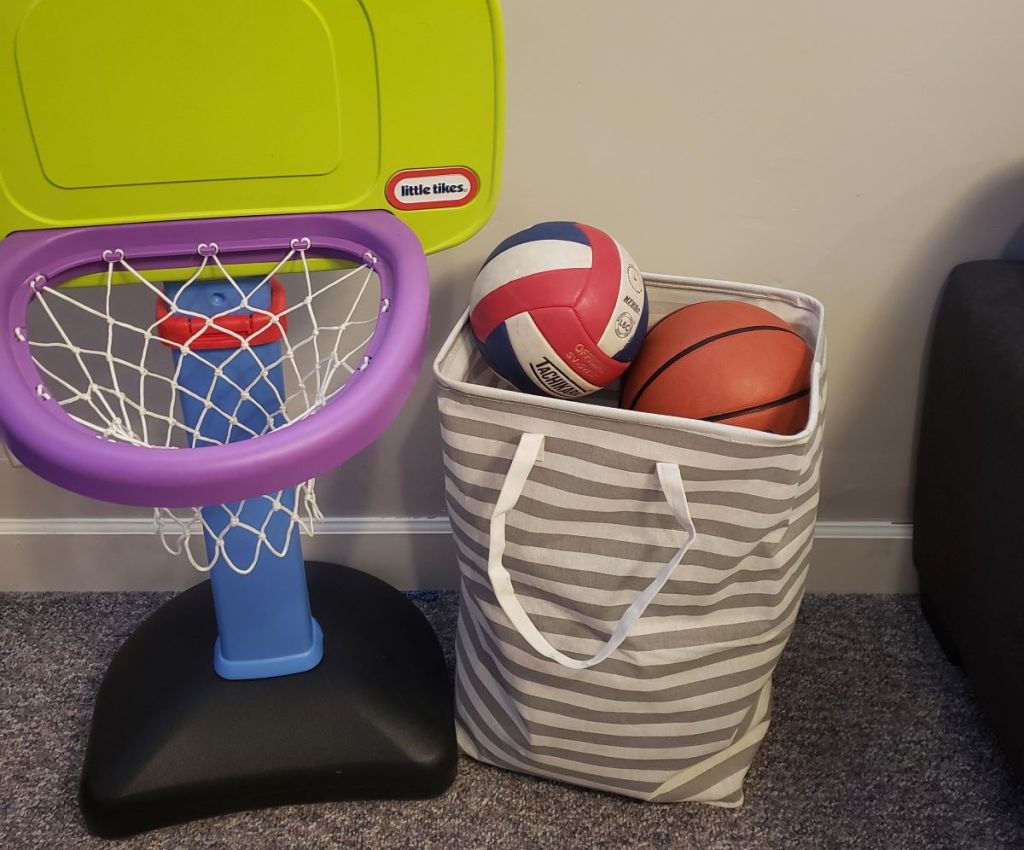 Hamper with a basketball and volleyball in it next to a toy basketball hoop