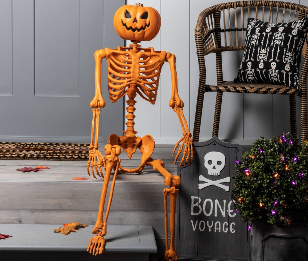 life size orange pumpkin head skeleton sitting on a porch with other Halloween decor