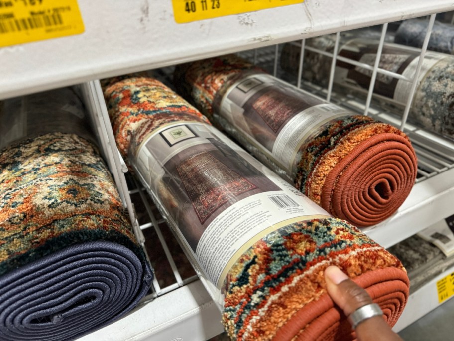 hand reaching for a rolled up runner rug with orange, black, brown and other colors on a store rack
